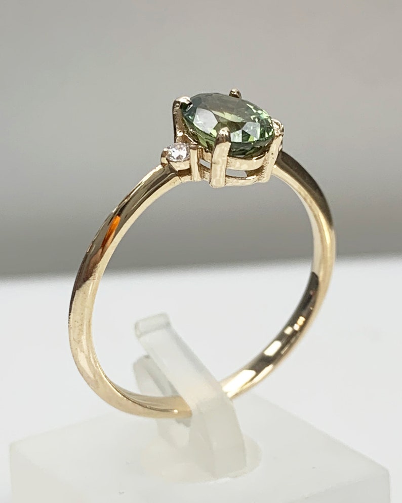 May include: A gold ring featuring a green oval gemstone and a smaller diamond accent. The ring is displayed on a white stand.