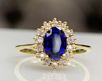 Op de afbeelding: Een gouden ring met een grote, ovaal geslepen, diepblauwe edelsteen, omringd door een halo van kleinere, heldere edelstenen. De ring heeft een dunne, gepolijste gouden band. De edelstenen reflecteren licht en schitteren.