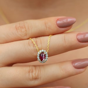 May include: A gold chain necklace with a red oval gemstone pendant surrounded by a halo of smaller clear gemstones.