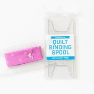 May include: A pink fabric spool with a white heart design and the text "The Original Quilt Binding Spool Containing One Spool" on a white background.