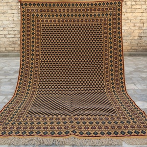 May include: A traditional hand-woven rug with a geometric pattern in shades of brown, beige, red, and blue. The rug has a fringe border and is displayed on a brick floor.
