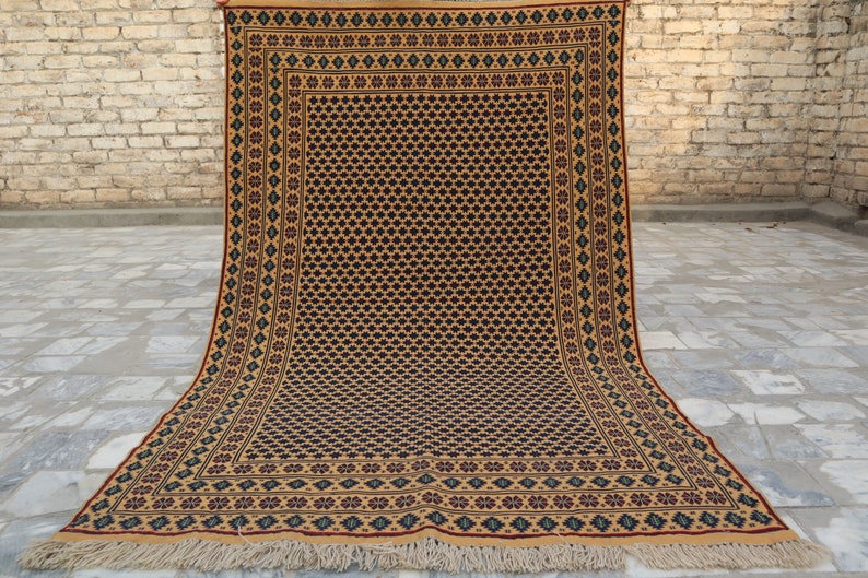 May include: A traditional hand-woven rug with a geometric pattern in shades of brown, beige, red, and blue. The rug has a fringe border and is displayed on a brick floor.