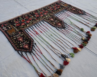 May include: A decorative textile with a black background, featuring red floral patterns and a brown deer motif. The textile has long, colorful tassels with various colored pom-poms at the ends. The edges are trimmed with a red and black border.