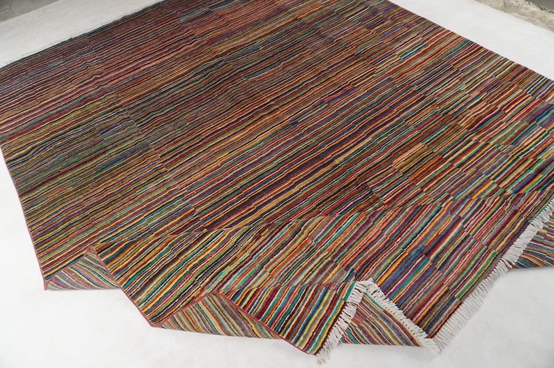 May include: A rectangular area rug featuring a dense pattern of thin, multicolored horizontal stripes in shades of red, blue, yellow, green, and orange. The rug has a woven texture and includes short white fringe detailing along the edges.