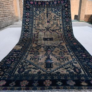 Peut inclure: Un long tapis rectangulaire avec un champ central beige et une bordure bleu fonc&eacute; complexe. Le tapis pr&eacute;sente un motif d&eacute;taill&eacute; avec des motifs floraux et g&eacute;om&eacute;triques, notamment des arbres stylis&eacute;s et des figures d'animaux. Le tapis est expos&eacute; &agrave; l'ext&eacute;rieur sur une surface blanche.