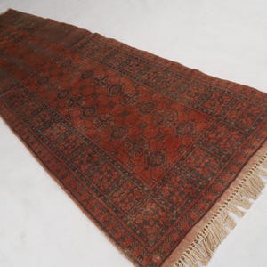 May include: A rectangular runner rug featuring a deep red base color with dark charcoal geometric patterns and repeating motifs. The rug is finished with light beige fringe tassels along the short ends, creating a textured border detail.