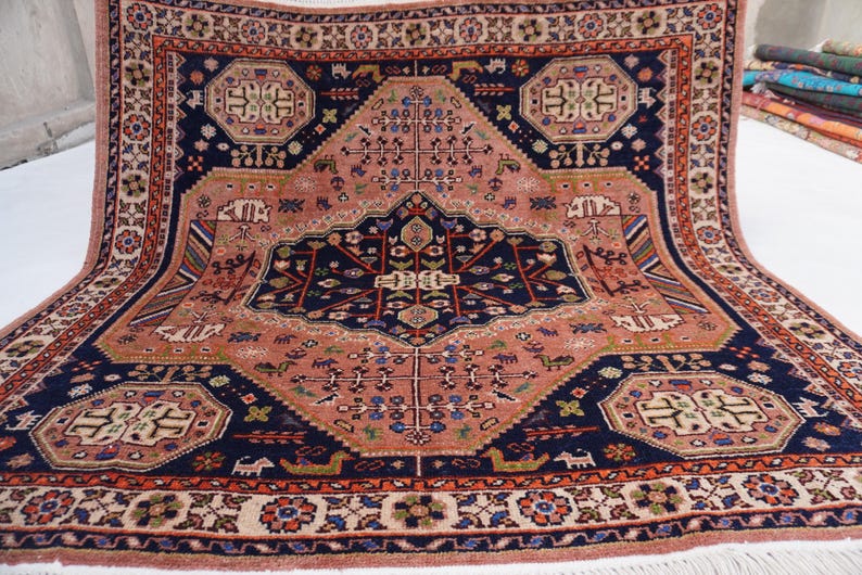 May include: A rectangular, hand-knotted rug with a central medallion design. The rug features a salmon-colored field with navy blue and rust-colored borders and accents. Intricate floral and geometric patterns adorn the rug's surface, with a fringe along the edges.