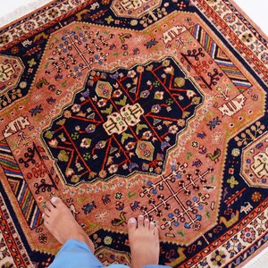 May include: A square, hand-knotted rug with a central navy blue medallion and a salmon-colored field. The rug features intricate floral and geometric patterns in red, green, and white, with a fringed border. The rug is a decorative floor covering.