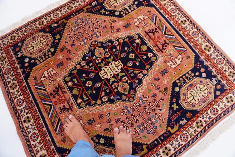 May include: A square, hand-knotted rug with a central navy blue medallion and a salmon-colored field. The rug features intricate floral and geometric patterns in red, green, and white, with a fringed border. The rug is a decorative floor covering.