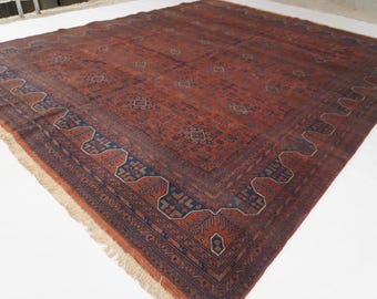 May include: A large square area rug featuring a deep terracotta red field with intricate dark blue geometric patterns and a wide border. The rug displays a repeating motif of small diamonds and stylized floral elements, finished with a light fringe.