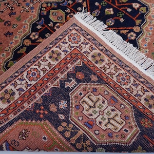 May include: A detailed view of a Persian rug with a complex geometric pattern. The rug features a combination of colors, including navy blue, peach, and orange. The edges are adorned with a fringe, and the design includes floral and geometric motifs.