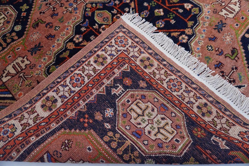 May include: A detailed view of a Persian rug with a complex geometric pattern. The rug features a combination of colors, including navy blue, peach, and orange. The edges are adorned with a fringe, and the design includes floral and geometric motifs.