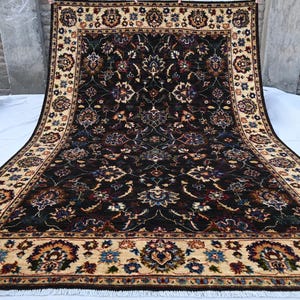 May include: A traditional Persian rug with a dark blue background and intricate floral patterns in shades of red, cream, and brown. The rug has a cream-coloured border with floral motifs.