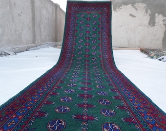 May include: A green and red patterned rug with a central design of repeating geometric shapes. The rug has a dark green field with red and blue accents. The border is a deep red with blue and gold details. The rug is displayed on a white surface.