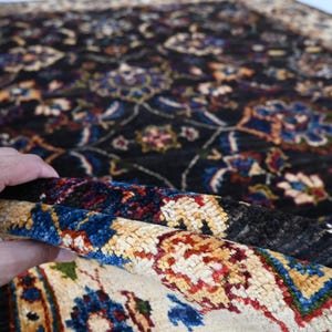 May include: A close-up of a colourful rug with intricate floral patterns in shades of blue, red, yellow, and white. The rug has a dark background and a hand is holding up a corner of the rug to reveal the lighter underside.
