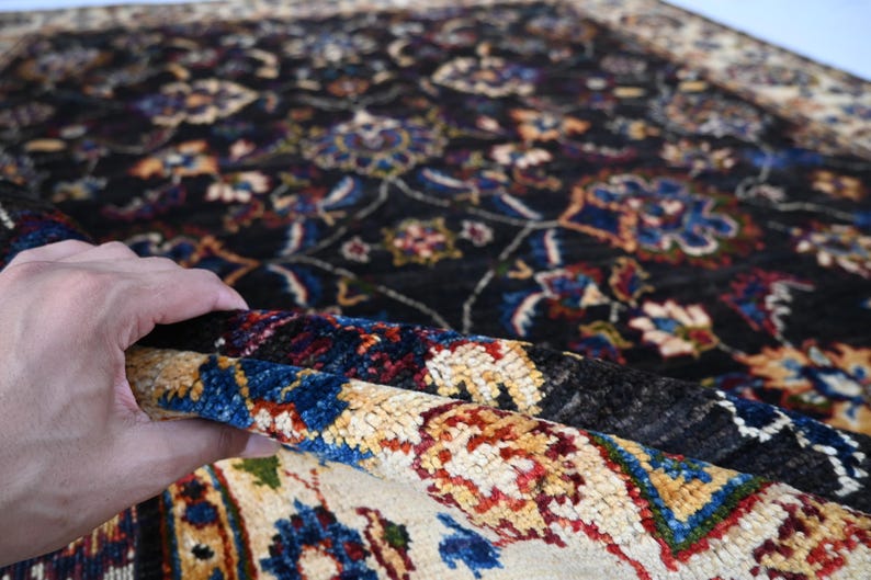 May include: A close-up of a colourful rug with intricate floral patterns in shades of blue, red, yellow, and white. The rug has a dark background and a hand is holding up a corner of the rug to reveal the lighter underside.
