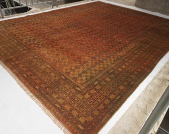 May include: A large, square area rug featuring a deep rust-orange and brown color palette. The rug displays a dense, repeating geometric pattern across its central field, framed by a wide, intricate border with detailed floral and linear motifs.