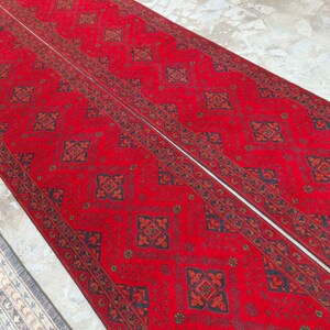 K&ouml;nnte beinhalten: Zwei lange, rote Teppiche mit Muster und einem wei&szlig;en Mittelstreifen. Die Teppiche weisen ein sich wiederholendes geometrisches Design in Schwarz und Braun auf. Die Teppiche liegen auf einer hellen Oberfl&auml;che, wobei ein Teil eines blau-wei&szlig;en Teppichs links sichtbar ist.