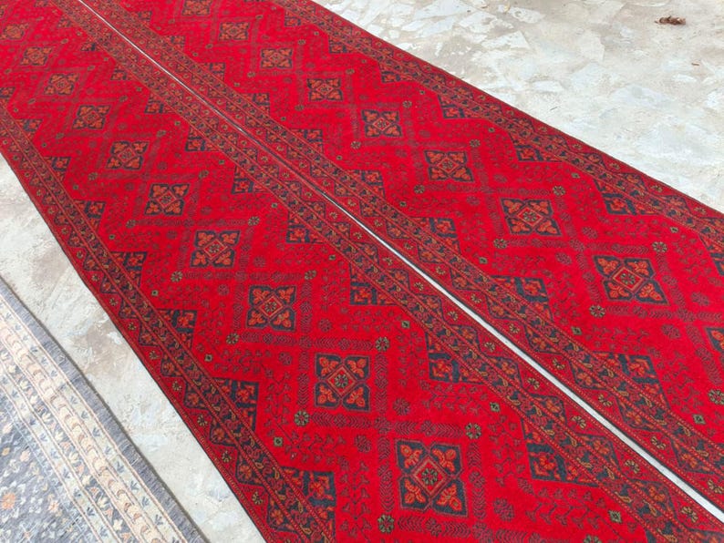 K&ouml;nnte beinhalten: Zwei lange, rote Teppiche mit Muster und einem wei&szlig;en Mittelstreifen. Die Teppiche weisen ein sich wiederholendes geometrisches Design in Schwarz und Braun auf. Die Teppiche liegen auf einer hellen Oberfl&auml;che, wobei ein Teil eines blau-wei&szlig;en Teppichs links sichtbar ist.