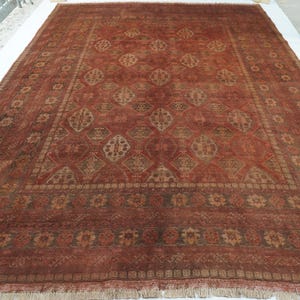 May include: A large rectangular area rug featuring a deep terracotta red base color with a repeating geometric diamond pattern in muted tan and brown tones. The rug has a wide decorative border with floral motifs and a short fringe along the edge.