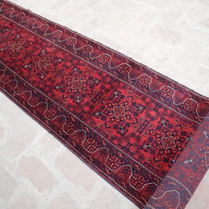 May include: A rolled-up, rectangular red runner rug with a detailed geometric pattern in navy blue and rust-orange. The rug's design includes repeating diamond and floral motifs, with a decorative border. The rug is placed on a light-colored brick surface.