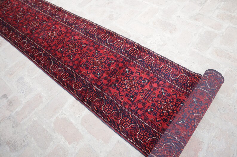 May include: A rolled-up, rectangular red runner rug with a detailed geometric pattern in navy blue and rust-orange. The rug's design includes repeating diamond and floral motifs, with a decorative border. The rug is placed on a light-colored brick surface.