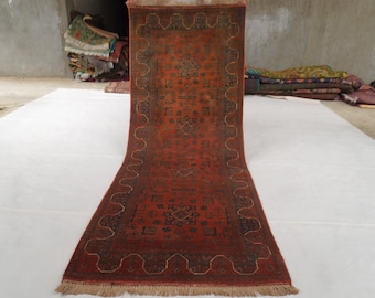 May include: A rectangular runner rug featuring a deep red background with intricate dark blue and black geometric patterns. The rug has a decorative scalloped border design and a light-colored fringe along the bottom edge, showcasing a detailed woven texture.