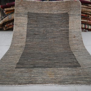 May include: A rectangular rug with a central dark gray square framed by a lighter gray border. The rug has a textured appearance, with visible horizontal lines. The edges are fringed. The rug is displayed on a light-colored surface.