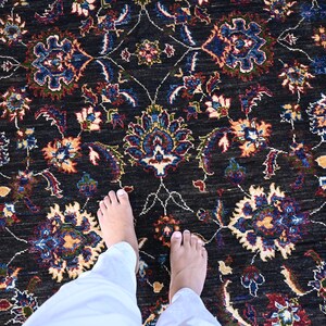 May include: A black rug with a floral pattern in shades of blue, red, and cream. The rug is shown with two bare feet standing on it.