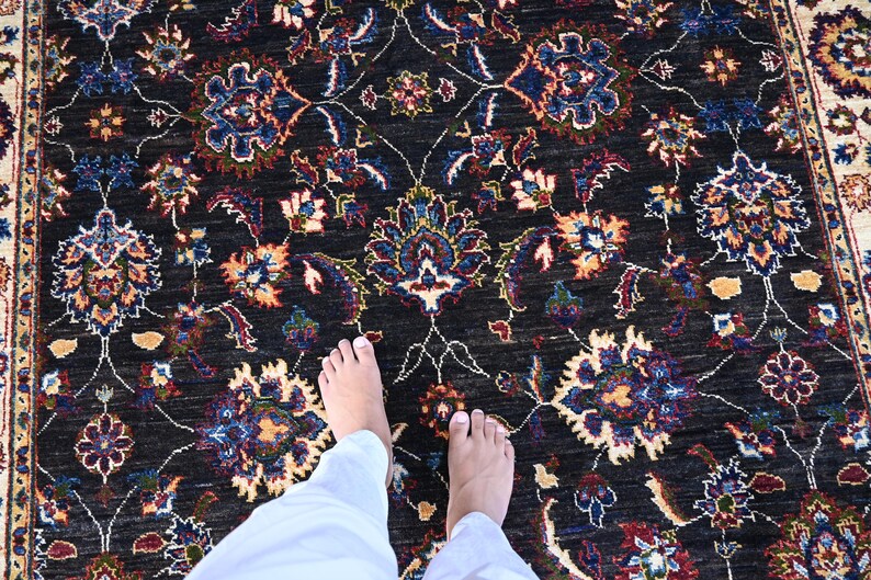 May include: A black rug with a floral pattern in shades of blue, red, and cream. The rug is shown with two bare feet standing on it.