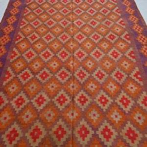 Op de afbeelding: Een geweven tapijt met een geometrisch patroon in tinten oranje, rood en paars. Het tapijt heeft een ruitpatroon met een rand van kleinere ruiten.