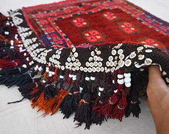 May include: A hand-woven rug with a deep red field and geometric patterns in shades of red, blue, and white. The rug features a detailed border and decorative tassels in various colors, including black, orange, and red.