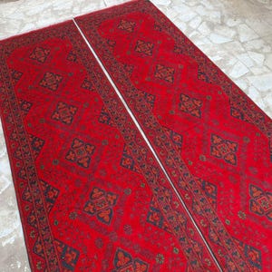 K&ouml;nnte beinhalten: Zwei lange, rechteckige rote Teppiche mit komplizierten geometrischen Mustern in Schwarz und Orange. Die Teppiche liegen nebeneinander und zeigen ein symmetrisches Design, ideal f&uuml;r einen Flur oder Eingangsbereich.