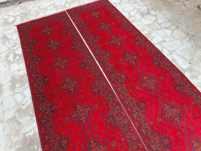 K&ouml;nnte beinhalten: Zwei lange, rechteckige rote Teppiche mit komplizierten geometrischen Mustern in Schwarz und Orange. Die Teppiche liegen nebeneinander und zeigen ein symmetrisches Design, ideal f&uuml;r einen Flur oder Eingangsbereich.