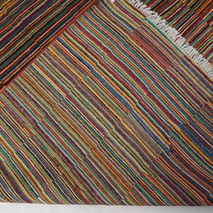 May include: A woven rug featuring a dense pattern of thin, multicolored stripes in shades of red, yellow, blue, green, and purple. The rug has a folded corner revealing a white fringe along the edge, showcasing a consistent striped texture.