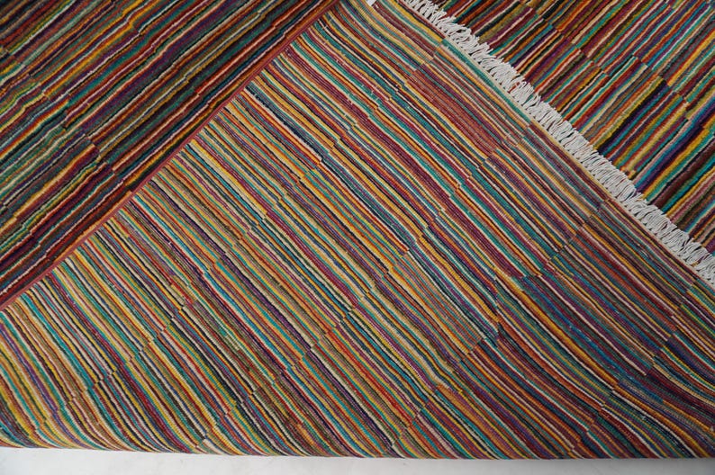 May include: A woven rug featuring a dense pattern of thin, multicolored stripes in shades of red, yellow, blue, green, and purple. The rug has a folded corner revealing a white fringe along the edge, showcasing a consistent striped texture.