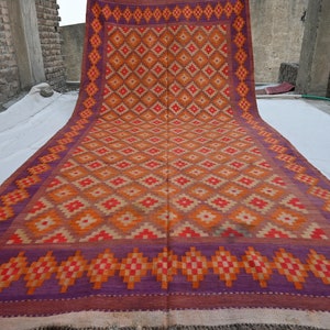 Op de afbeelding: Een groot, handgeweven tapijt met een geometrisch patroon in tinten oranje, rood en paars. Het tapijt ligt op een wit oppervlak en heeft een franje rand.