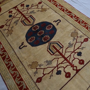 May include: A beige rug with a red, blue, and green geometric pattern. The rug features a central motif of a stylized tree with branches and fruit. The border of the rug is a repeating pattern of red, green, and beige.