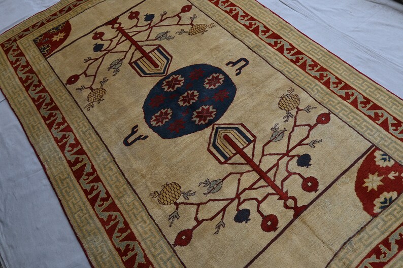 May include: A beige rug with a red, blue, and green geometric pattern. The rug features a central motif of a stylized tree with branches and fruit. The border of the rug is a repeating pattern of red, green, and beige.