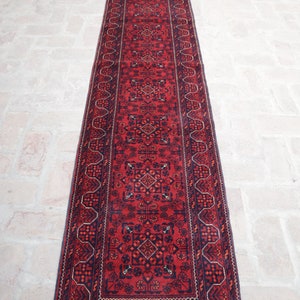 May include: A long, rectangular red rug with an intricate geometric pattern. The rug features a central field of red with navy blue and dark orange accents. The border is navy blue with a repeating design. The rug has a fringed edge.