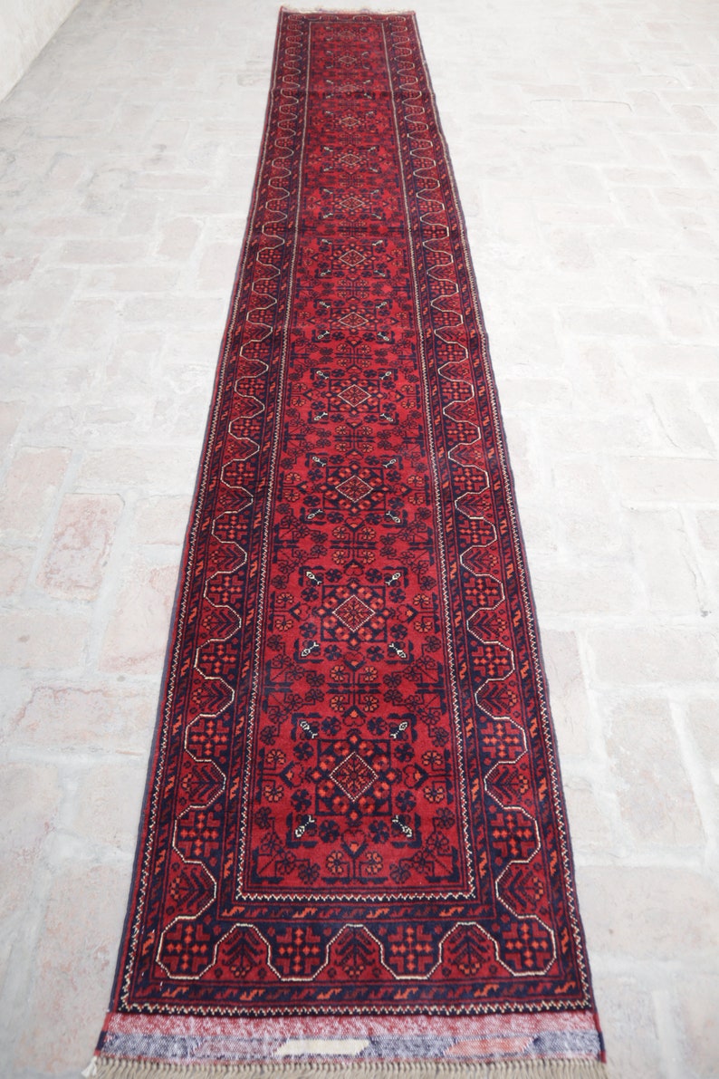May include: A long, rectangular red rug with an intricate geometric pattern. The rug features a central field of red with navy blue and dark orange accents. The border is navy blue with a repeating design. The rug has a fringed edge.