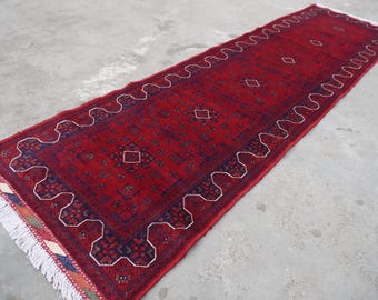 May include: A long, rectangular red rug with a detailed pattern. The rug features a central field of red with geometric designs and a border with a white, wavy pattern. The rug has a fringe on both ends.