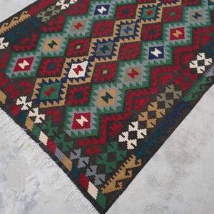 May include: A colorful rug with a geometric pattern in shades of red, green, blue, brown, and white. The rug has a fringe border.
