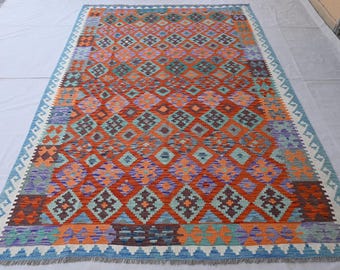 May include: A rectangular rug with a geometric pattern in shades of orange, blue, purple, and green. The rug features a diamond and square design, with a blue and white border. The rug is made of woven material.
