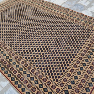 May include: A beige rug with a black and blue geometric pattern. The rug has a red and green border with a fringe.