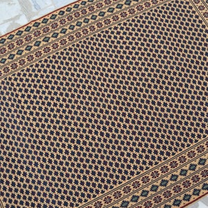 May include: A beige rug with a black and white geometric pattern. The rug has a border with a floral design in shades of red, green, and blue.