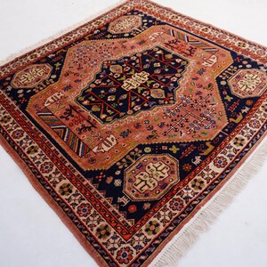 May include: Square, hand-knotted rug with a detailed, geometric design. The rug features a central navy blue medallion surrounded by a peach field and a border of red and cream. The rug has a fringe on the edges.