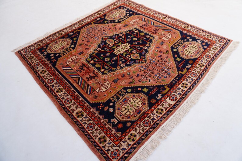 May include: Square, hand-knotted rug with a detailed, geometric design. The rug features a central navy blue medallion surrounded by a peach field and a border of red and cream. The rug has a fringe on the edges.