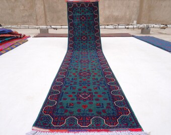 May include: A long, rectangular rug with a teal background and intricate red and navy blue geometric patterns. The rug has a decorative border and fringed edges. Other rugs are visible in the background.