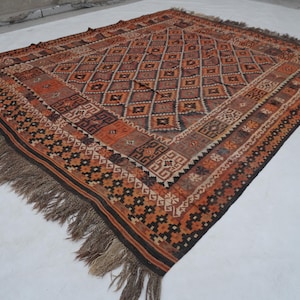 May include: A rectangular, hand-woven rug with a geometric diamond pattern in shades of brown, orange, and beige. The rug has a dark brown border and fringe along the edges. The rug is likely made of wool.